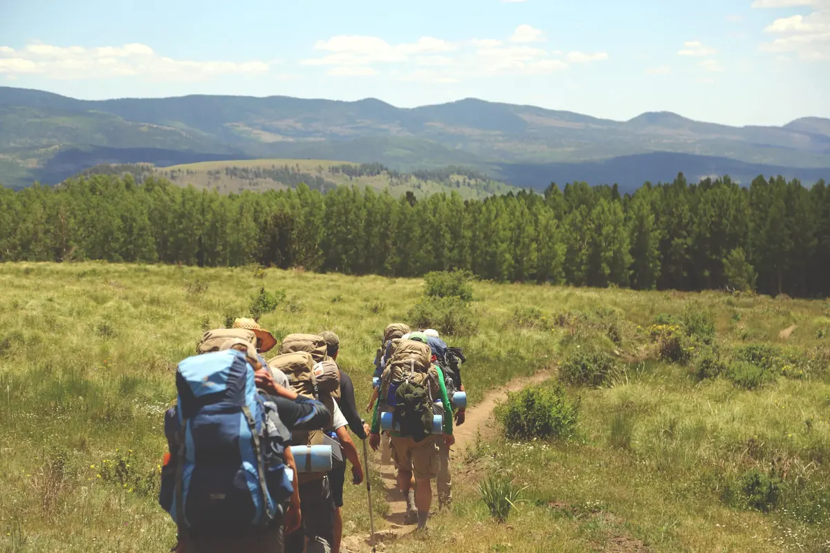 A small group of hikers with backpacks walking single file along a dirt path through a grassy meadow towards pine forest and distant hills