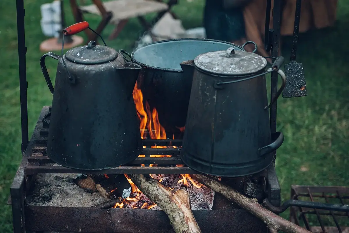 Blackened enamel coffee pots and a cooking pot on a steel grate over a wood fire, on grass
