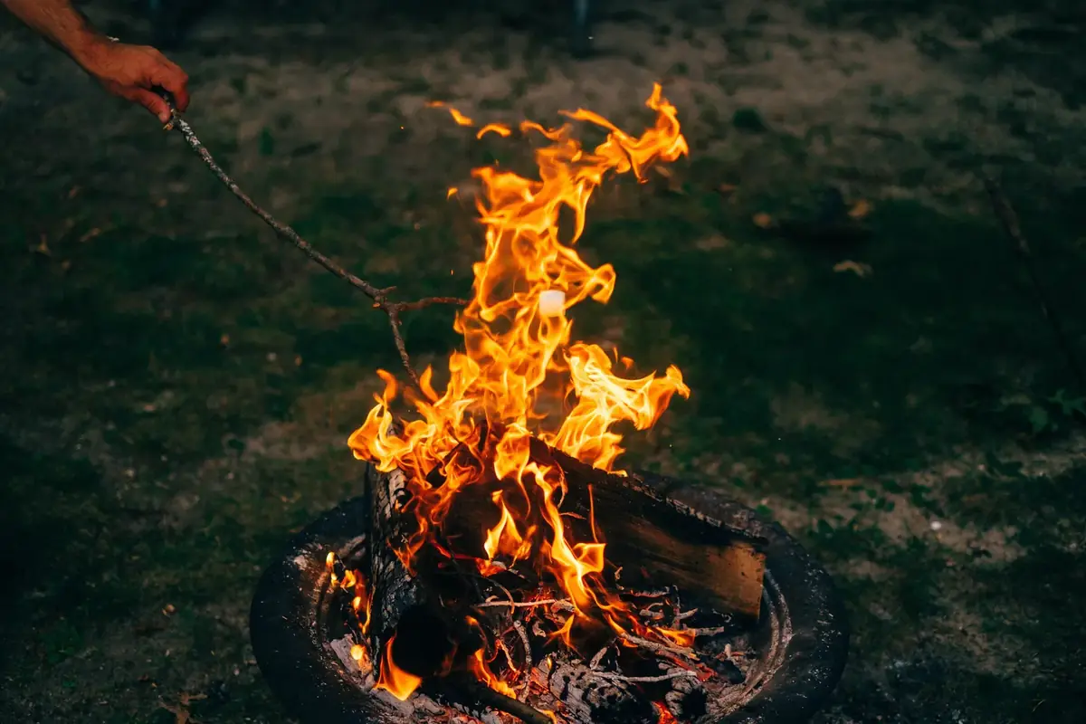 A wood fire burning in a round metal fire ring on grass at dusk, with someone tending it with a stick