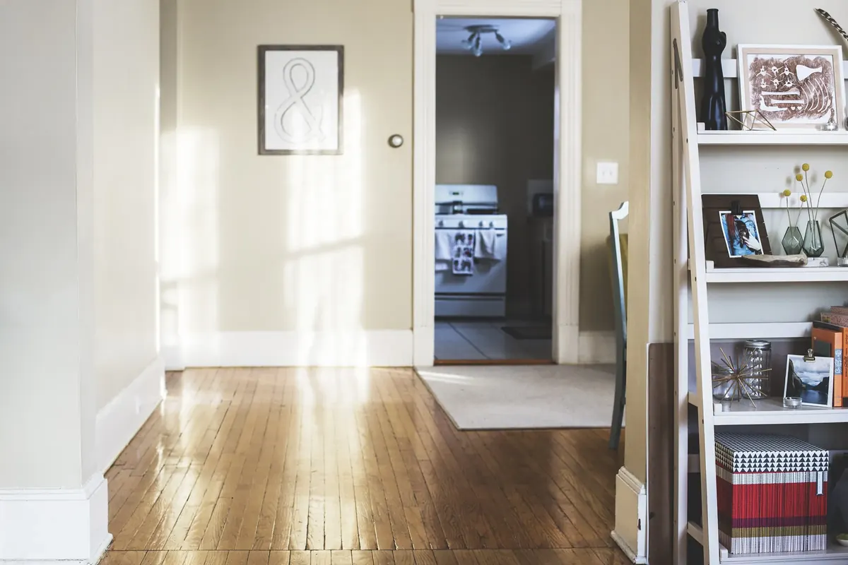 An empty, sunlit room with a polished wooden floor, white walls and an open door to a kitchen, with a white ladder shelf of books and pictures at one side