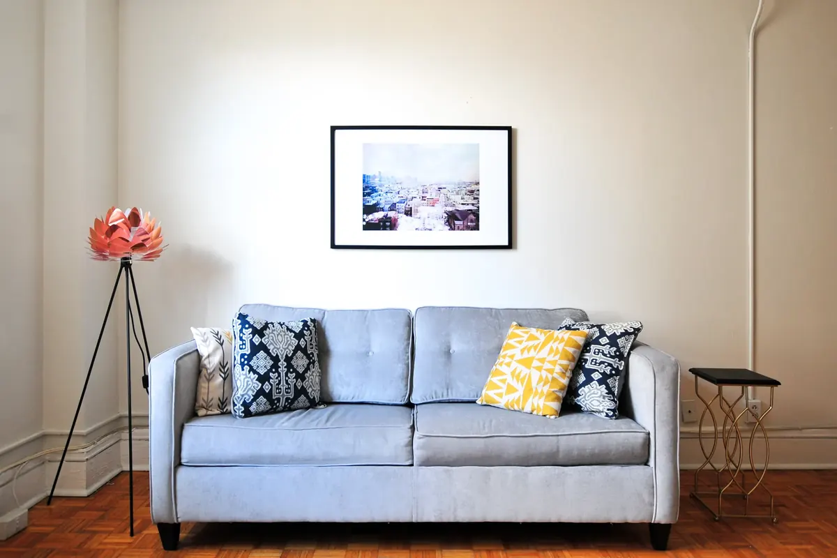 A small pale blue sofa with patterned cushions, a framed print and a tripod floor lamp against a plain white wall above a wooden floor