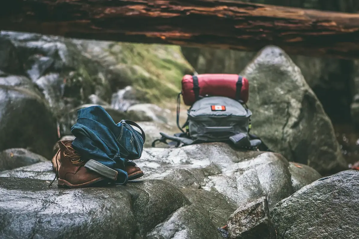 A small backpack with a red sleeping bag strapped on top, beside leather boots and a folded denim jacket on wet rocks