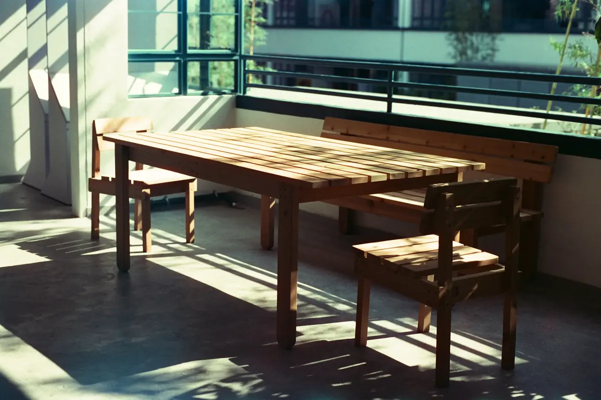 A plain wooden table and chairs in strong sunlight by a long window, with shadows of the window bars across the floor
